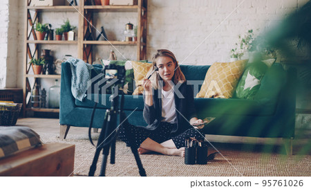 Popular female blogger is recording video about make-up and beauty sitting on floor at home and using camera on tripod. Girl is showing professional cosmetics kit and using brush. 95761026