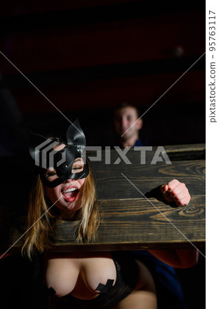 nipple tapes. A woman is imprisoned in a wooden pillory during sexual games. BDSM Erotic fantasies. Subordination. Toys for adults. The mask of the cat. The man in the shirt dominates. 95763117