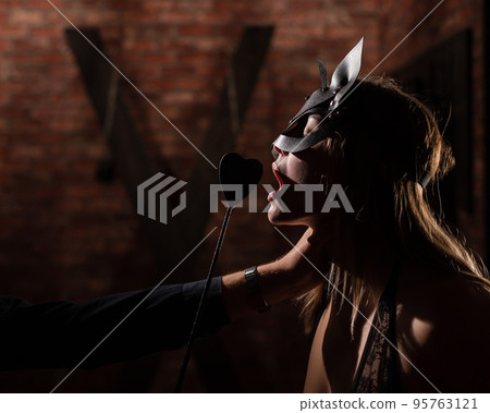 A man in a shirt holds his mistress by the neck. A woman in a leather cat mask bites a leather whip. BDSM Games for adults. Sensual lips. A man in a shirt holds his mistress by the neck. A woman in a leather cat mask bites a leather whip. BDSM Games for adults. Sensual lips. 95763121