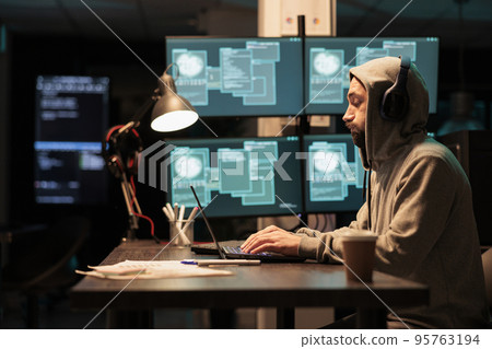 Male criminal trying to break database server, using virus to code malware and steal big data at night. Working with multiple monitors and hacking information system, wearing hood. Male criminal trying to break database server, using virus to code malware and steal big data at night. Working with multiple monitors and hacking information system, wearing hood. 95763194