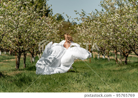A woman in a white dress runs through a blossoming cherry orchard. The long dress flies to the sides, the bride runs rejoicing in life. 95765769