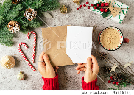 Desktop of woman hands in red sweater holding blank paper and envelope . Flat lay of gray background with cup of coffee and Christmas decoration. Top view mock up and copy space for text 95766134