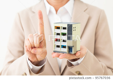 A middle-aged woman in a suit holding a model of an apartment house and raising her index finger 95767036