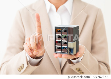 A middle-aged woman in a suit holding a model of an apartment house and raising her index finger 95767138