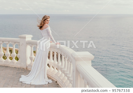 Romantic bride, blond girl in white wedding dress with open shoulders posing on open terrace with backdrop of the sea and rocks. Stylish young woman standing on terrace and looking on ocean at sunset Romantic bride, blond girl in white wedding dress with open shoulders posing on open terrace with backdrop of the sea and rocks. Stylish young woman standing on terrace and looking on ocean at sunset 95767338