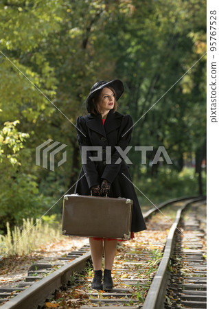 Madame in clothes of 30-40s style mysteriously looks away. Woman in black hat and coat holds retro valise.Vertical frame Madame in clothes of 30-40s style mysteriously looks away. Woman in black hat and coat holds retro valise.Vertical frame 95767528