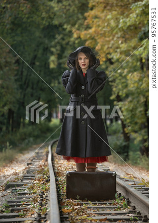 Middle-aged woman in retro black coat, broad-brim hat stands near vintage suitcase on railroad tracks. Vertical frame 95767531
