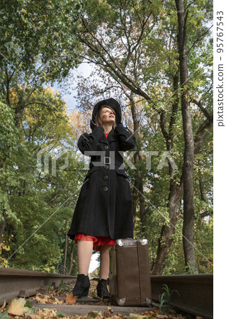 Middle-aged woman in long coat and black hat with brims looks up at sky. Madam stands on rails with suitcase in forest Middle-aged woman in long coat and black hat with brims looks up at sky. Madam stands on rails with suitcase in forest 95767543