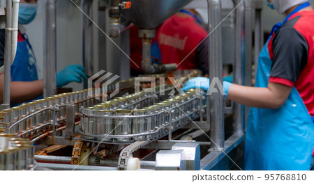 Canned fish factory. Food industry.  Sardines in red tomato sauce in tinned cans on conveyor belt at food factory. Blur workers working in food processing production line. Food manufacturing industry. 95768810