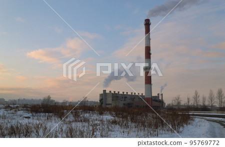 Boiler room with a tall striped smoking chimney in the middle of a winter evening landscape Boiler room with a tall striped smoking chimney in the middle of a winter evening landscape 95769772