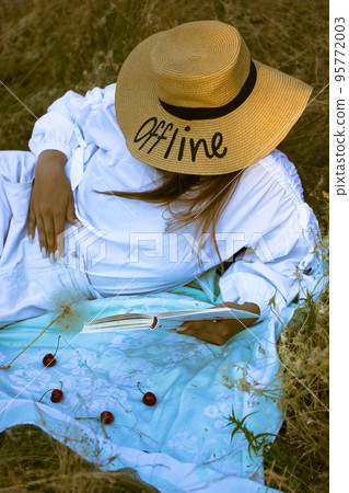 Mysterious tourist girl, a young stylishly dressed woman in a straw hat with the text Offline lying on a plaid reading a paper book. Picnic in nature in the summer, autumn on vacation Literature books Mysterious tourist girl, a young stylishly dressed woman in a straw hat with the text Offline lying on a plaid reading a paper book. Picnic in nature in the summer, autumn on vacation Literature books 95772003