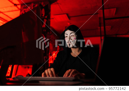 Unrecognizable hacker man wearing anonymous mask and sweatshirt typing on wireless keyboard breaking password using laptop and desktop pc, sitting at desk in dark basement room with blue neon lights. Unrecognizable hacker man wearing anonymous mask and sweatshirt typing on wireless keyboard breaking password using laptop and desktop pc, sitting at desk in dark basement room with blue neon lights. 95772142