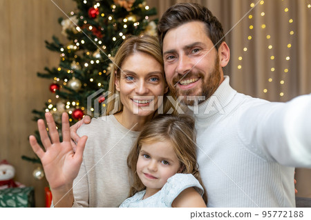 Festive portrait of a young happy family near the Christmas tree at home. Mom, dad and daughter take a selfie, look at the camera, smile, wave. 95772188