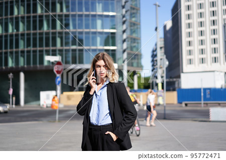 Portrait of businesswoman in suit, standing on street with mobile phone, on call with someone, smiling while having telephone conversation Portrait of businesswoman in suit, standing on street with mobile phone, on call with someone, smiling while having telephone conversation 95772471