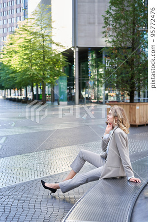 Vertical shot of young business woman in beige suit, office worker taking break outside near office building Vertical shot of young business woman in beige suit, office worker taking break outside near office building 95772476