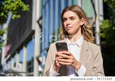 Portrait of stylish businesswoman in corporate outfit, holding smartphone, standing on street and using mobile phone in hands 95772493