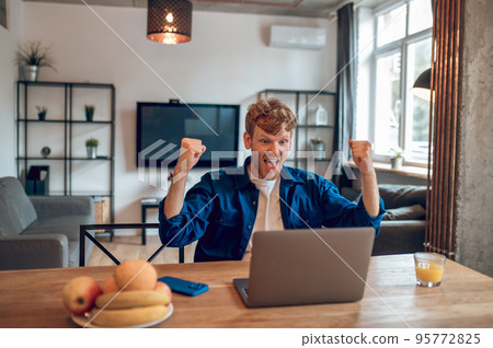 Man sitting in the home office at the laptop and looking excited Man sitting in the home office at the laptop and looking excited 95772825