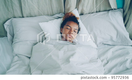 Young African American woman is sleeping in comfortable bed under warm blanket having rest on beautiful linen. Comfort, resting people and bedroom concept. Young African American woman is sleeping in comfortable bed under warm blanket having rest on beautiful linen. Comfort, resting people and bedroom concept. 95772936