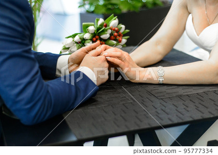 Happy bride and groom on their wedding. An amazing bride bouquet white tulips, the new husband and wife at a table holding each other's hands Happy bride and groom on their wedding. An amazing bride bouquet white tulips, the new husband and wife at a table holding each other's hands 95777334