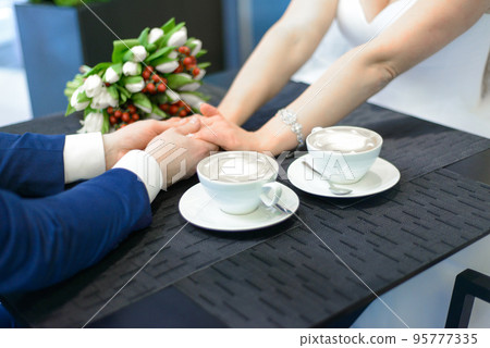Happy bride and groom on their wedding. An amazing bride bouquet white tulips, the new husband and wife at the table ready to drink coffee Happy bride and groom on their wedding. An amazing bride bouquet white tulips, the new husband and wife at the table ready to drink coffee 95777335
