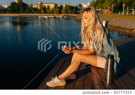 Happy relaxed blonde young woman holding cup with hot coffee from thermos sitting on beach by wooden pier on sunny summer morning. Beautiful female traveler enjoying with nature drinking tea over lake 95777353