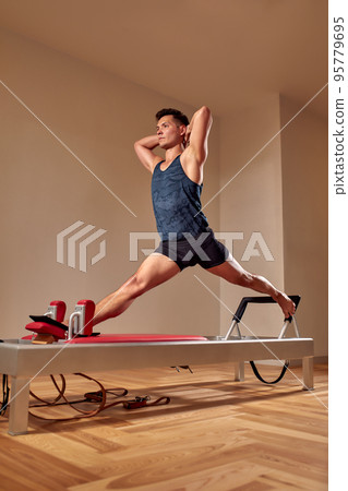 Side view of barefoot male athlete lying on pilates reformer and performing abs exercise during fitness workout. Pilates man concept. Side view of barefoot male athlete lying on pilates reformer and performing abs exercise during fitness workout. Pilates man concept. 95779695
