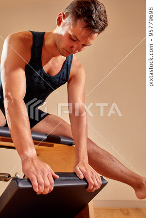 Handsome pilates male instructor performing stretching balance fitness exercise on small barrel equipment, at the pilates studio modern onterior indoor. Handsome pilates male instructor performing stretching balance fitness exercise on small barrel equipment, at the pilates studio modern onterior indoor. 95779698