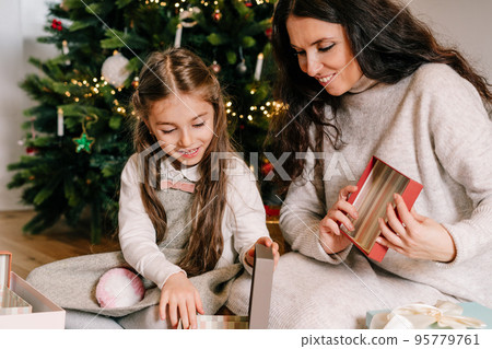 Excited daughter and mother sitting at home near beautiful decorated Christmas tree and enjoying opening their presents. Family holidays. Festive christmas mood. Selective focus. 95779761
