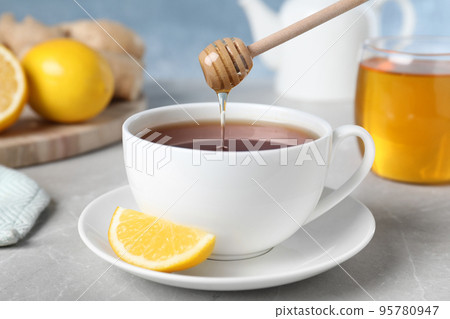 Honey pouring into cup with tea from wooden dipper on light grey marble table, closeup 95780947