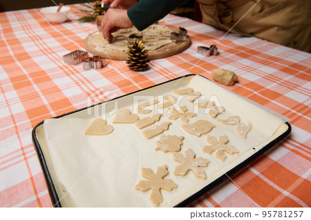 A baking sheet with gingerbread cookies from various shape on the parchment, on the background of a housewife confectioner cutting molds out of dough. Baking cookies for Christmas holidays. New Year A baking sheet with gingerbread cookies from various shape on the parchment, on the background of a housewife confectioner cutting molds out of dough. Baking cookies for Christmas holidays. New Year 95781257