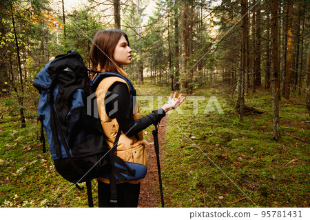 European young girl tourist with large backpack walks through forest with compass and walking sticks European young girl tourist with large backpack walks through forest with compass and walking sticks 95781431