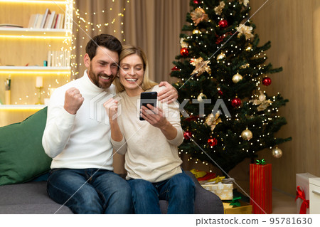 Happy christmas couple man and woman using smartphone smiling and happy celebrating victory discount and shopping in online store near christmas tree Happy christmas couple man and woman using smartphone smiling and happy celebrating victory discount and shopping in online store near christmas tree 95781630