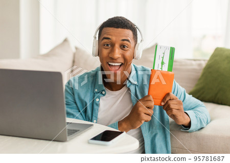Glad excited young african american guy in wireless headphones with passport, air tickets, near computer 95781687