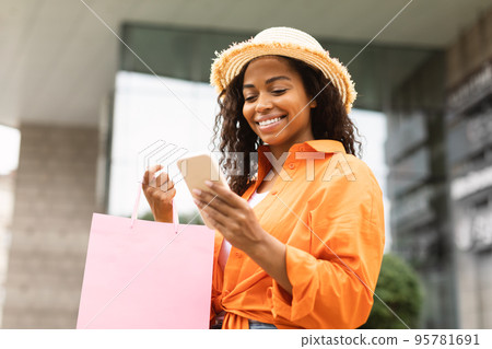 Happy young pretty african american woman in casual, hat with many bags with purchases typing on smartphone 95781691