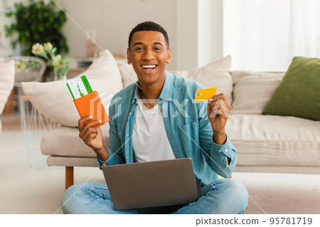 Cheerful millennial african american guy with laptop shows passport, air ticket and credit card, rejoices in trip 95781719
