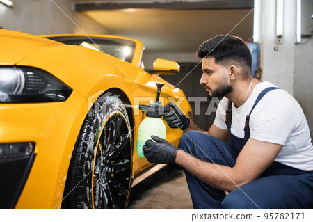 Indoor shot of male worker in overalls, washing the car wheels rims Indoor shot of male worker in overalls, washing the car wheels rims 95782171