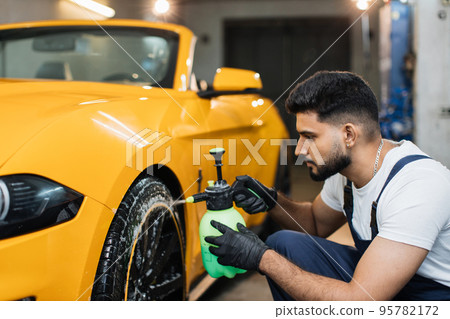 Indoor shot of male worker in overalls, washing the car wheels rims 95782172