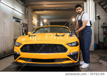 Full length portrait of young handsome man, worker of auto detailing service, holds orbital polisher Full length portrait of young handsome man, worker of auto detailing service, holds orbital polisher 95782189