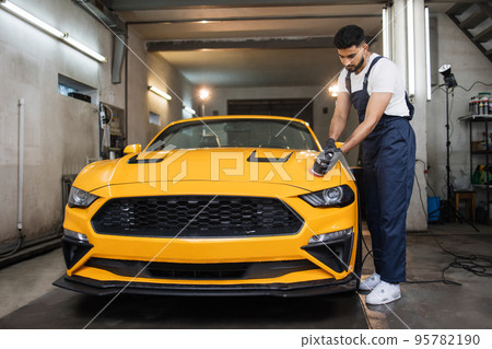 Full length portrait of young handsome man, worker of auto detailing service, holds orbital polisher 95782190