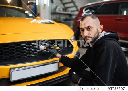 Male worker in gloves washes car radiator grille of luxury yellow car with special brush and soap Male worker in gloves washes car radiator grille of luxury yellow car with special brush and soap 95782307