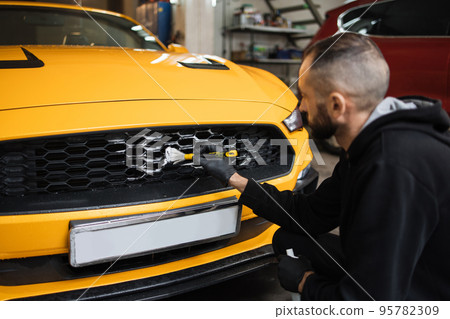 Male worker in gloves washes car radiator grille of luxury yellow car with special brush and soap Male worker in gloves washes car radiator grille of luxury yellow car with special brush and soap 95782309