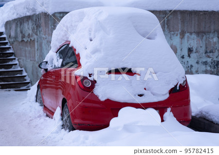 Red car in the parking space. The car is covered in snow. Huge snowdrift on the car Red car in the parking space. The car is covered in snow. Huge snowdrift on the car 95782415