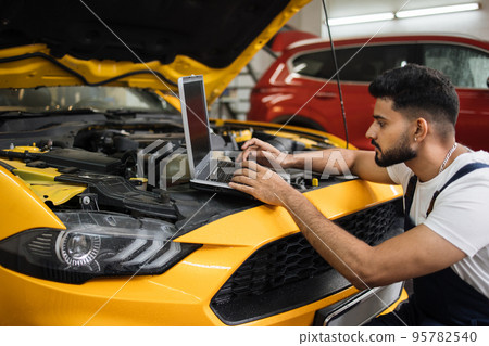 Young bearded man mechanic using laptop for auto diagnostics of sport yellow car engine Young bearded man mechanic using laptop for auto diagnostics of sport yellow car engine 95782540