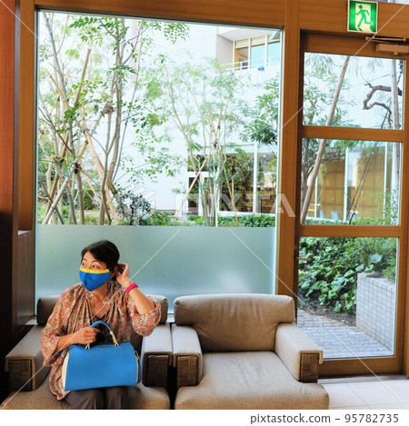 A woman in a mask sitting on a sofa by a bright window A woman in a mask sitting on a sofa by a bright window 95782735