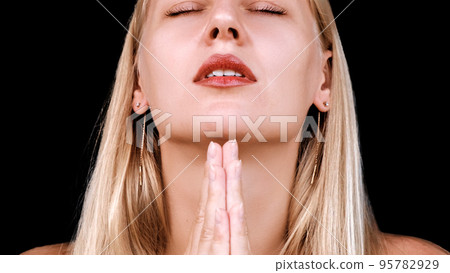 Woman prays to God. Portrait of young blonde girl folding her palms together in prayer pose. Woman says a prayer with her head raised and her eyes closed on a black background. Religion, faith Woman prays to God. Portrait of young blonde girl folding her palms together in prayer pose. Woman says a prayer with her head raised and her eyes closed on a black background. Religion, faith 95782929