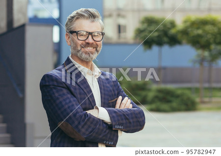 Portrait of mature gray-haired investor, senior man in glasses looking at camera and gesturing, businessman with crossed arms outside office building in business suit, successful boss banker. Portrait of mature gray-haired investor, senior man in glasses looking at camera and gesturing, businessman with crossed arms outside office building in business suit, successful boss banker. 95782947