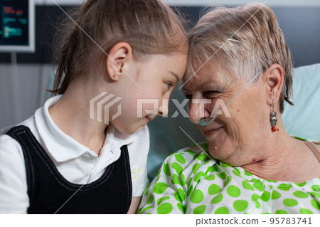 Elderly woman lying in hospital bed with breathing aids showing affection to little girl. Grandmother and granddaughter bumping heads in love at medical clinic for elderly people recovery room. 95783741