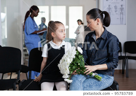 Mom, daughter waiting to see hospitalized family member in hospital reception. Girl, young woman in hospital visiting area. Relatives bringing flowers to hospital patient. 95783789