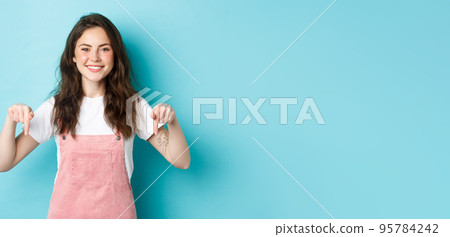 Place your logo here. Cheerful brunette woman pointing fingers down and smiling at camera, recommending product, showing sale promo offer, standing against blue background Place your logo here. Cheerful brunette woman pointing fingers down and smiling at camera, recommending product, showing sale promo offer, standing against blue background 95784242