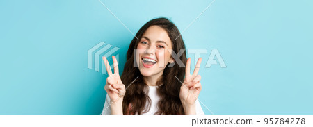 Young positive modern girl sending good vibes only, showing v-sign and smiling happy, standing against blue background 95784278